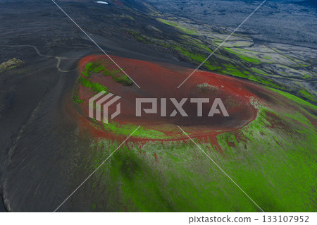 Aerial image of a volcanic crater in Iceland with vivid red and green tones, surrounded by black terrain, a dirt road, and distant rugged landscapes. 133107952
