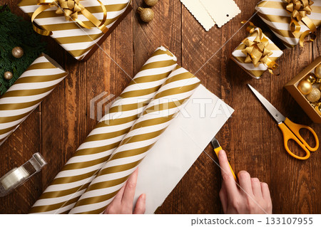 Present preparation. Hands cut gold and white striped wrapping paper with a stationery knife on a brown wooden table. 133107955