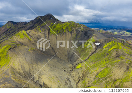 Aerial view of a volcanic crater in Iceland, featuring green moss, dark soil, a patch of snow, rolling hills, and distant plains under a cloudy sky. 133107965