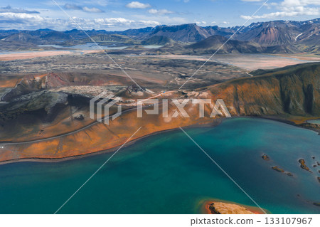 Aerial view of a turquoise lake surrounded by orange and brown hills in Iceland's Landmannalaugar region, with snow capped mountains in the background. 133107967