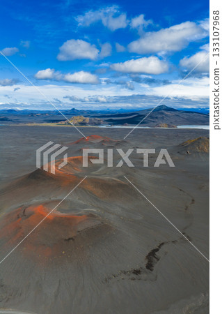 Aerial view of reddish orange volcanic craters in Iceland, surrounded by dark terrain. Distant mountains, a lake, and a blue sky with clouds are visible. 133107968