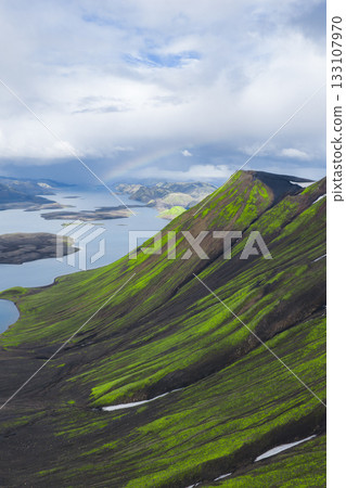 Aerial view of a green and black volcanic mountain slope with snow patches, a serene lake with islands, and dramatic clouds in Iceland. 133107970
