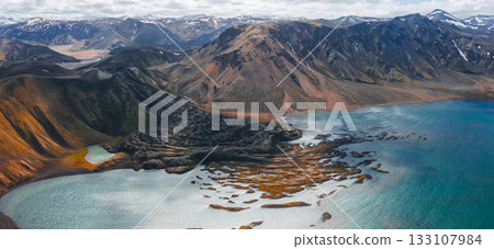 Aerial view of a volcanic mountain with black lava flow, turquoise lake, colorful rhyolite hills, snow capped peaks, and distant valleys in Iceland. 133107984