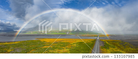 A vibrant rainbow spans an Icelandic landscape with a straight road, lush vegetation, and a waterfall cascading down a cliff under a partly cloudy sky. 133107985
