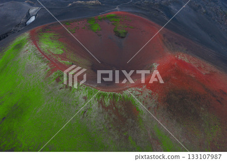 A striking aerial view of a volcanic crater in Iceland, featuring red tones in the interior, green moss on slopes, and a path in the background. 133107987