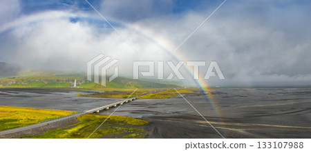 A rainbow arches over black sand and braided rivers in Iceland. A bridge spans the landscape, with a distant waterfall cascading down a misty cliff. A rainbow arches over black sand and braided rivers in Iceland. A bridge spans the landscape, with a distant waterfall cascading down a misty cliff. 133107988
