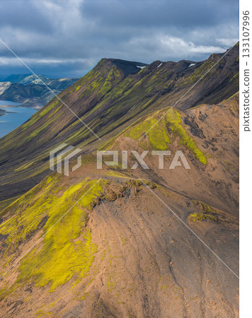 Aerial view of Iceland's vibrant landscape with green moss covered mountains, dark volcanic soil, a serene lake, and cloudy skies in the background. Aerial view of Iceland's vibrant landscape with green moss covered mountains, dark volcanic soil, a serene lake, and cloudy skies in the background. 133107996