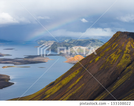 Aerial view of a moss in Iceland covered mountain slope, a serene lake with islands, rainbow tinted mountains, and a faint rainbow under a cloudy sky. Aerial view of a moss in Iceland covered mountain slope, a serene lake with islands, rainbow tinted mountains, and a faint rainbow under a cloudy sky. 133108022