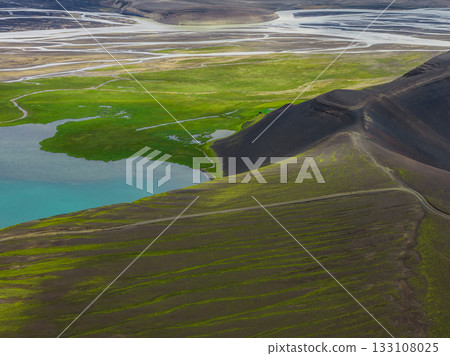 A vibrant turquoise lake surrounded by green vegetation, dark rugged hills with trails, and braided rivers flowing across a flat plain in Iceland. 133108025