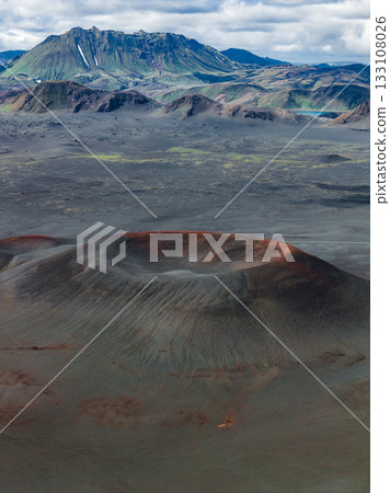 A reddish brown volcanic crater in Iceland surrounded by barren terrain, with rugged mountains, green vegetation, and snow under a partly cloudy sky. A reddish brown volcanic crater in Iceland surrounded by barren terrain, with rugged mountains, green vegetation, and snow under a partly cloudy sky. 133108026