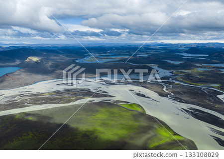 Aerial perspective of braided rivers crossing a volcanic plain with green moss covered hills, distant lakes, craters, and mountains under a cloudy sky. 133108029