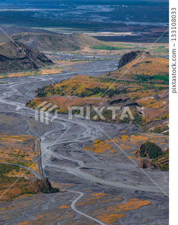 Braided river flows through green and yellow vegetation with volcanic soil, framed by rolling hills, rocky outcrops, and distant flatlands in Iceland. 133108033