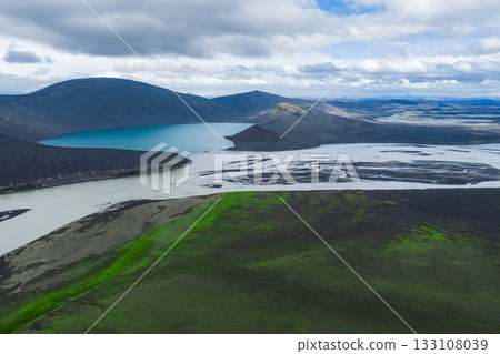 A turquoise crater lake sits amid dark volcanic terrain in Iceland, with a winding river, green moss patches, and distant hills under a partly cloudy sky. A turquoise crater lake sits amid dark volcanic terrain in Iceland, with a winding river, green moss patches, and distant hills under a partly cloudy sky. 133108039