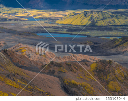 Aerial view of Iceland's rugged landscape with moss covered craters, vibrant hills, a serene blue lake, and contrasting green and yellow vegetation. Aerial view of Iceland's rugged landscape with moss covered craters, vibrant hills, a serene blue lake, and contrasting green and yellow vegetation. 133108044