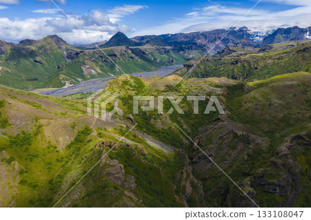 Aerial view of a green valley in Iceland with deep ravines, rolling hills, and a braided river. Snow capped peaks and glaciers are visible under a blue sky. Aerial view of a green valley in Iceland with deep ravines, rolling hills, and a braided river. Snow capped peaks and glaciers are visible under a blue sky. 133108047