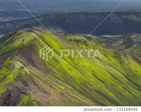 Vibrant green moss covered hills with dark volcanic rock, a dramatic ridge, and a flat plain in Iceland's unique volcanic terrain. 133108048