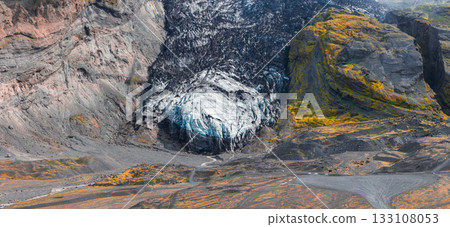 A glacier in Iceland surrounded by steep cliffs with volcanic rock, yellow moss, and a winding path leading to the icy blue and white glacier. 133108053