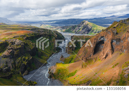 Mulagljufur Canyon in Iceland features dramatic cliffs, green vegetation, a winding river, and a weathered rock formation under a partly cloudy sky. 133108056