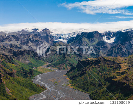 A winding river in Iceland flows through a lush green valley surrounded by cliffs and jagged mountains, with snow capped peaks and glaciers in the background. 133108059
