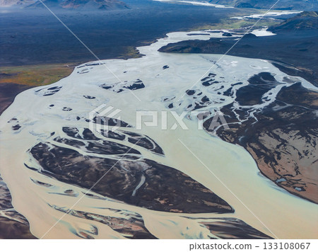 Aerial view of a pale blue glacial river weaving through dark volcanic sands, with distant hills and mountains in Iceland's rugged landscape. 133108067