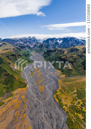 Aerial view of a braided river in a lush green valley with volcanic sediment, golden hues, snow capped peaks, and glaciers in Iceland's landscape. 133108068