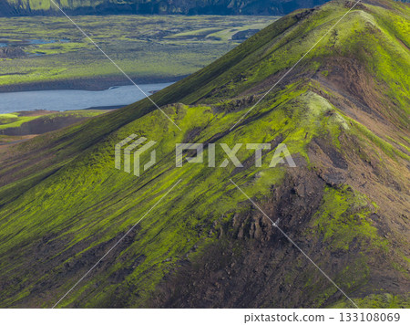A green moss covered mountain ridge with sharp peaks contrasts with volcanic plains and a winding body of water under soft natural light in Iceland. 133108069