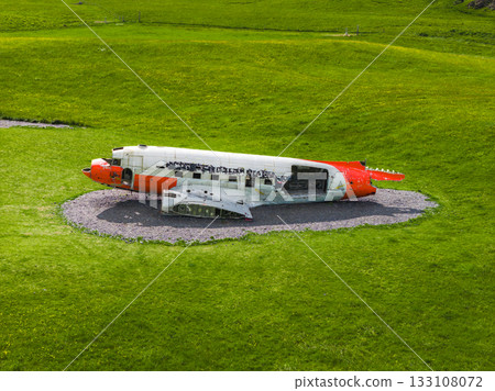 Partially intact Abandoned U.S. Navy Douglas C 117D aircraft fuselage in white and orange lies on gravel, surrounded by vibrant green grass in an open area, likely in Iceland. 133108072