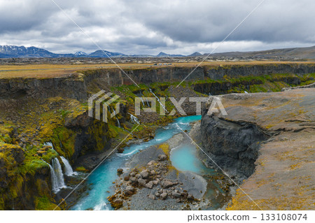 Turquoise river winds through Sigoldugljufur Canyon in Iceland, surrounded by mossy cliffs, cascading waterfalls, and distant snow capped mountains. 133108074