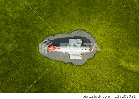 An aerial view of a weathered red and white Abandoned U.S. Navy Douglas C 117D aircraft on gravel, surrounded by vibrant green grass in Iceland's dramatic natural landscape. An aerial view of a weathered red and white Abandoned U.S. Navy Douglas C 117D aircraft on gravel, surrounded by vibrant green grass in Iceland's dramatic natural landscape. 133108081
