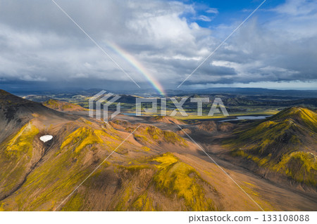Aerial view of Iceland's Landmannalaugar region with vibrant rhyolite mountains, green moss, a rainbow, snow patches, valleys, and scattered lakes. 133108088