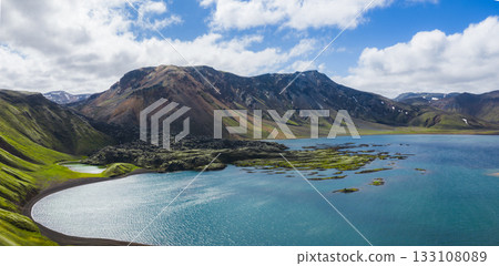 A vivid blue lake surrounded by green moss covered hills and multicolored mountains with snow capped peaks under a partly cloudy sky in Iceland. 133108089