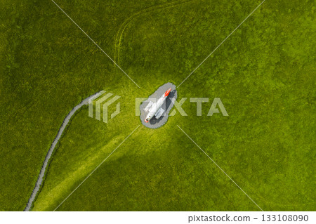 An aerial view shows a white Abandoned U.S. Navy Douglas C 117D aircraft monument with a red nose on a circular gravel area, surrounded by a vast green field and a narrow path in Iceland 133108090