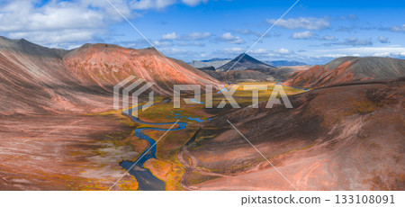 Aerial view of colorful rhyolite mountains, a winding river, and lush green valley under a bright blue sky with a triangular peak in the background. 133108091