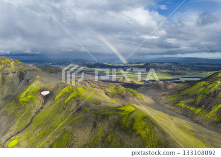 Aerial view of Iceland's moss covered hills, valleys, and volcanic terrain with a rainbow, rivers, lakes, and dramatic clouds in the highlands. 133108092