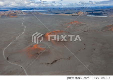 Aerial view of a barren volcanic terrain in Iceland, featuring reddish orange craters, a winding dirt road, distant mountains, and scattered clouds. Aerial view of a barren volcanic terrain in Iceland, featuring reddish orange craters, a winding dirt road, distant mountains, and scattered clouds. 133108095