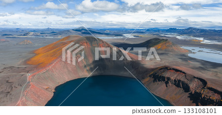 A volcanic crater lake in Iceland with steep red and orange walls, deep blue water, rocky plains, winding rivers, and distant mountains under cloudy skies. 133108101