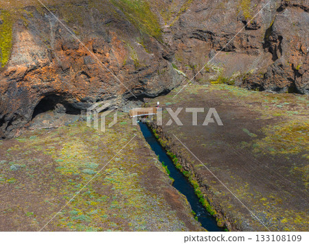 A vivid blue stream flows through a moss covered canyon with steep rocky cliffs. A small wooden bridge spans the stream, adding a human element. 133108109