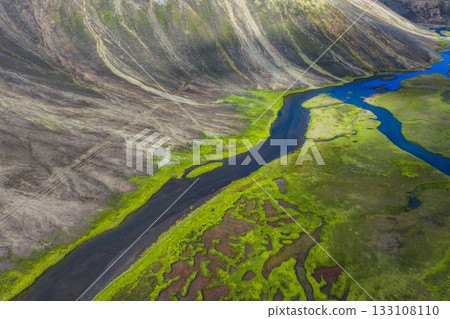 Aerial view of a winding river flowing through a green valley in Iceland, surrounded by steep, striated hills with earthy tones and moss patches. 133108110