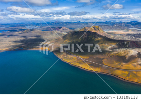 Aerial view of a volcanic landscape in Iceland featuring a crater like hill, vivid blue lake, winding road, rugged terrain, and distant mountains. 133108118