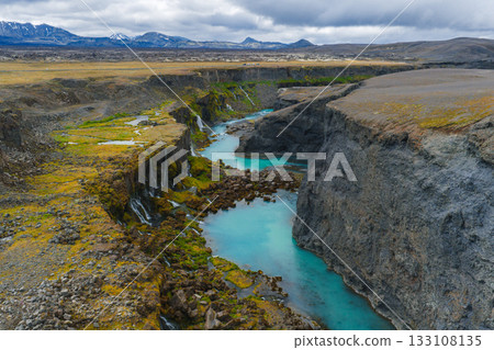 Sigoldugljufur Canyon features moss covered cliffs, cascading waterfalls, a turquoise river, rocky terrain, and distant snow capped mountains under cloudy skies. 133108135