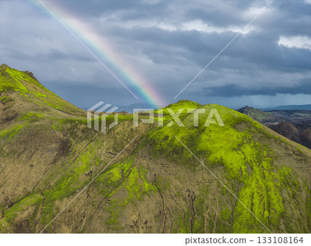 A vibrant green moss covered hill in Iceland with a dramatic rainbow arching across a cloudy sky, set against rugged volcanic terrain. A vibrant green moss covered hill in Iceland with a dramatic rainbow arching across a cloudy sky, set against rugged volcanic terrain. 133108164