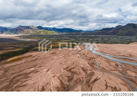 Aerial view of a braided river system in Iceland on a reddish brown plain, surrounded by rugged, multicolored mountains with green vegetation under a cloudy sky. 133108166