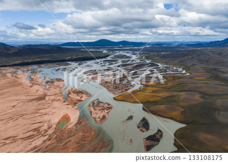 Aerial view of intertwining braided river channels in Iceland, surrounded by reddish brown sediment, green moss, volcanic soil, and distant mountains. 133108175