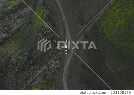 Aerial image of Iceland's volcanic landscape with green moss patches, featuring three vehicles at a dirt road intersection in a vast, isolated area. 133108179