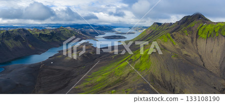 Aerial view of elongated lakes amid volcanic terrain and green moss covered mountains in Iceland, framed by dramatic cloud cover and sharp ridges. 133108190