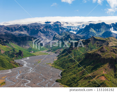 Aerial view of Mulagljufur Canyon in Iceland, featuring lush green cliffs, a braided river, volcanic soil, a glacier, and rugged mountains. 133108192