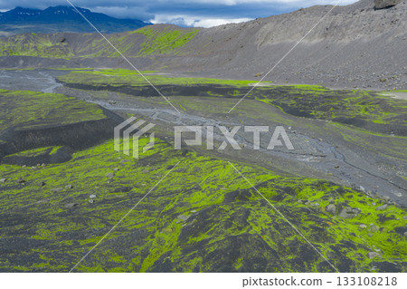 A winding river flows through dark volcanic terrain with vibrant green moss, set against a snowy mountain under a cloudy sky in Iceland. A winding river flows through dark volcanic terrain with vibrant green moss, set against a snowy mountain under a cloudy sky in Iceland. 133108218