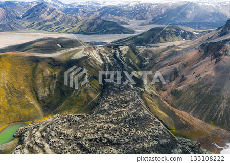 Aerial view of colorful rhyolite mountains in Landmannalaugar, Iceland, with a dark lava flow, patches of snow, and a small green lake in the foreground. 133108222