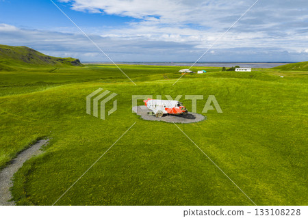 A red and white Abandoned U.S. Navy Douglas C 117D aircraft sits on a circular gravel patch in a green field, surrounded by grassy hills, small buildings in Iceland 133108228