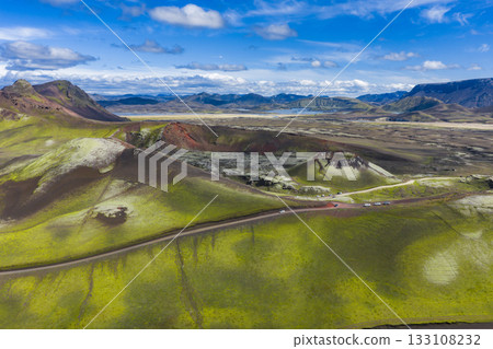 Aerial view of a volcanic crater surrounded by green moss covered hills, dark volcanic soil, a winding road, distant mountains, and a serene lake. Aerial view of a volcanic crater surrounded by green moss covered hills, dark volcanic soil, a winding road, distant mountains, and a serene lake. 133108232